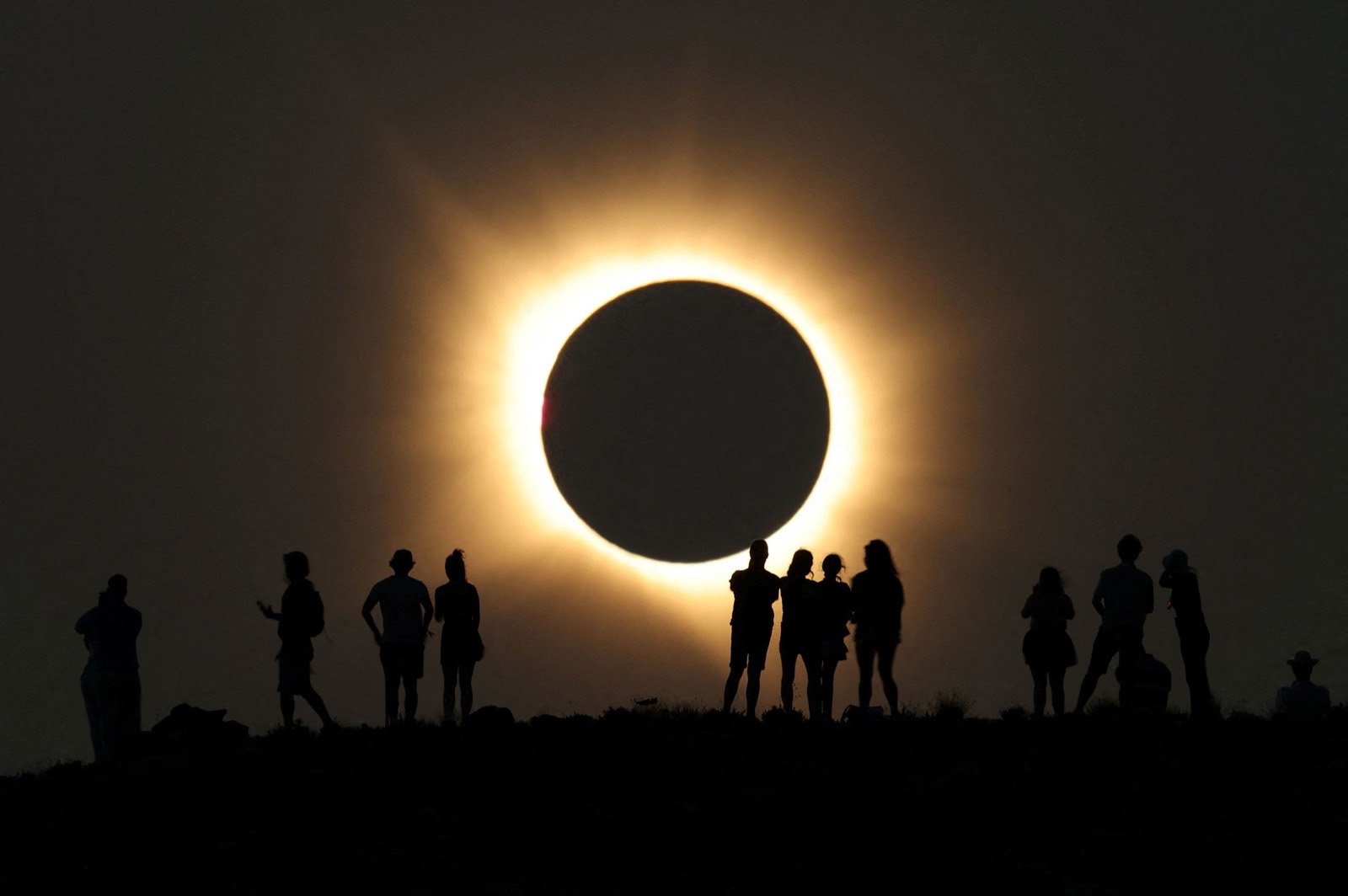 People silhouetted on a ridge watching a total solar eclipse, with the Moon covering the Sun and a bright corona